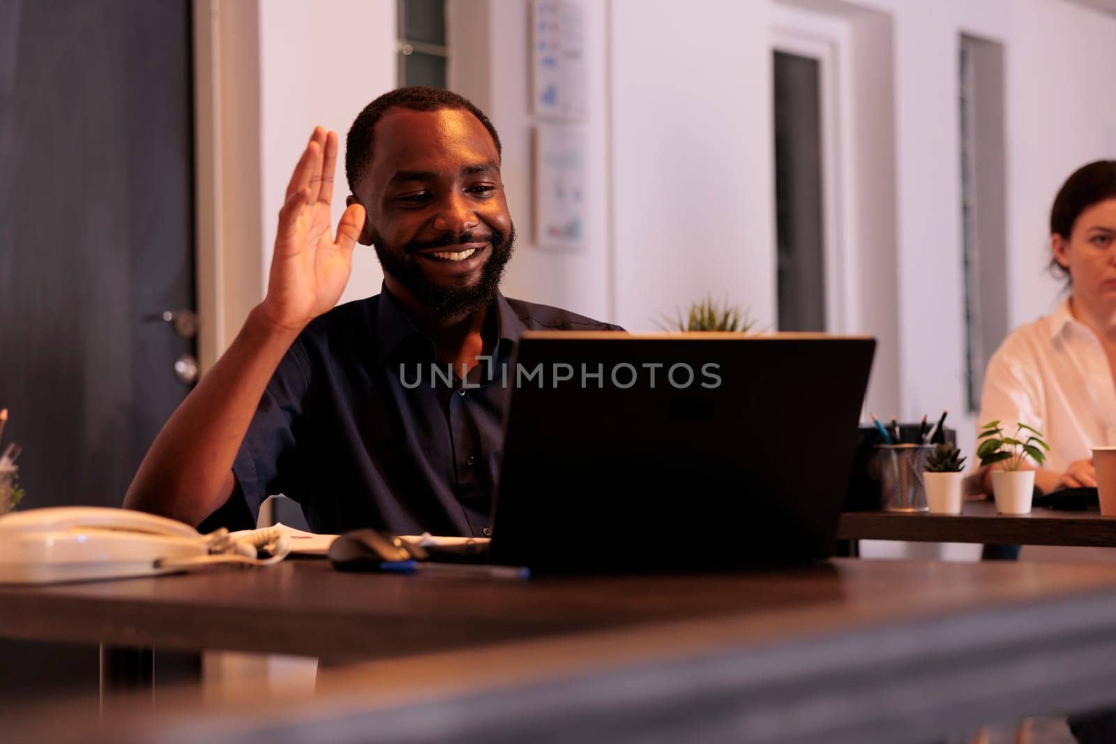 Smiling employee talking on videoconference with teamlead, waving hi, corporate worker answering teleconference call in office at night. African american man remote conversation in coworking space