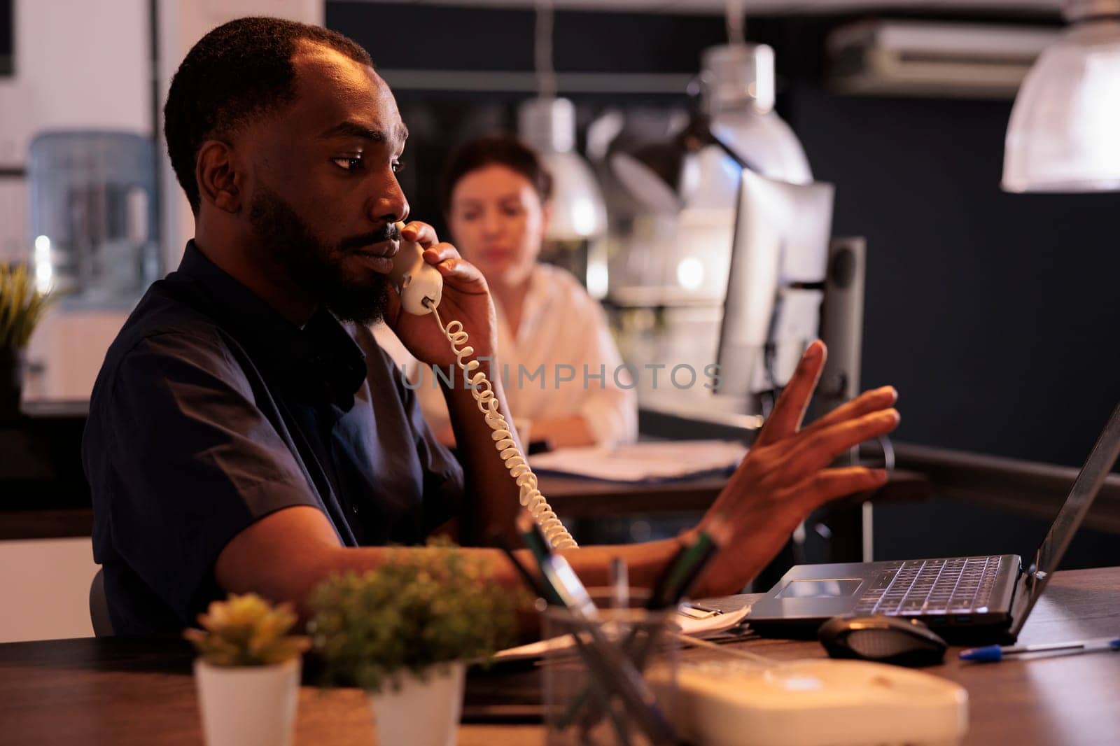 Corporate worker discussing business plan with teamlead on landline phone, employee answering call in office at night. African american man communication in coworking space with sunset light