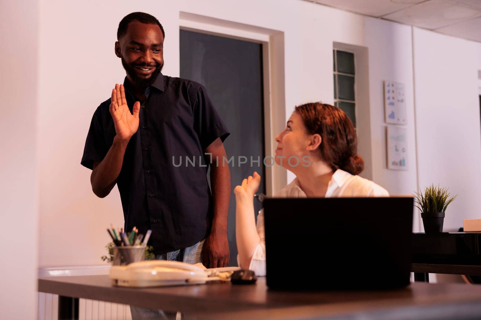 Office manager greeting new colleague, waving hi in coworking space, woman working on laptop, talking with coworker. Smiling african american man meeting employee, workers communication