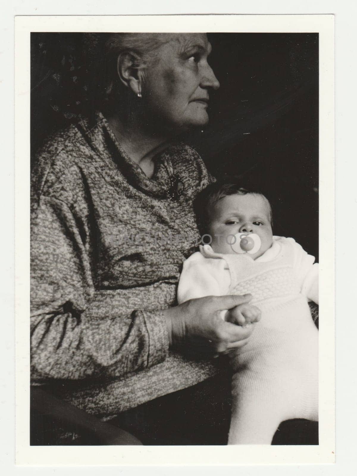 Vintage photo shows grandmother cradles todlder with dummy. Retro black and white photography. Circa 1970s. by roman_nerud