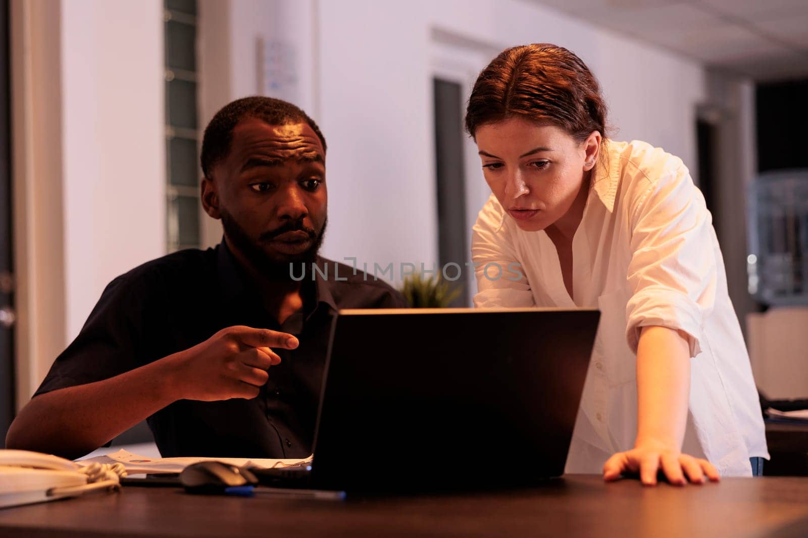 Colleagues working together on laptop in office, planning company strategy, discussing report. Corporate workers checking analytics research results at workplace desk in coworking space at night