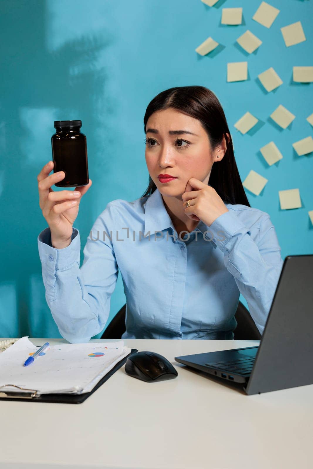Professional asian startup employee with headache looking at bottle of pills analyzing the components of contains. Woman at office desk looking doubtfully at a container of tablets.