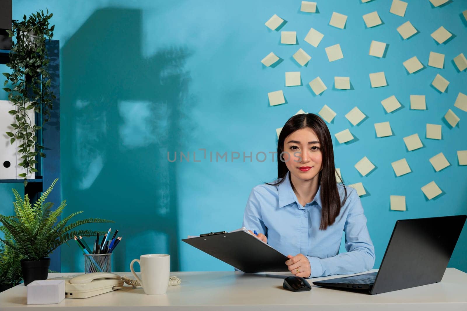 Woman sitting at desk and taking notes of office supplies replenishment inventory. Company female employee in charge of purchasing department with laptop and coffee cup on desk.