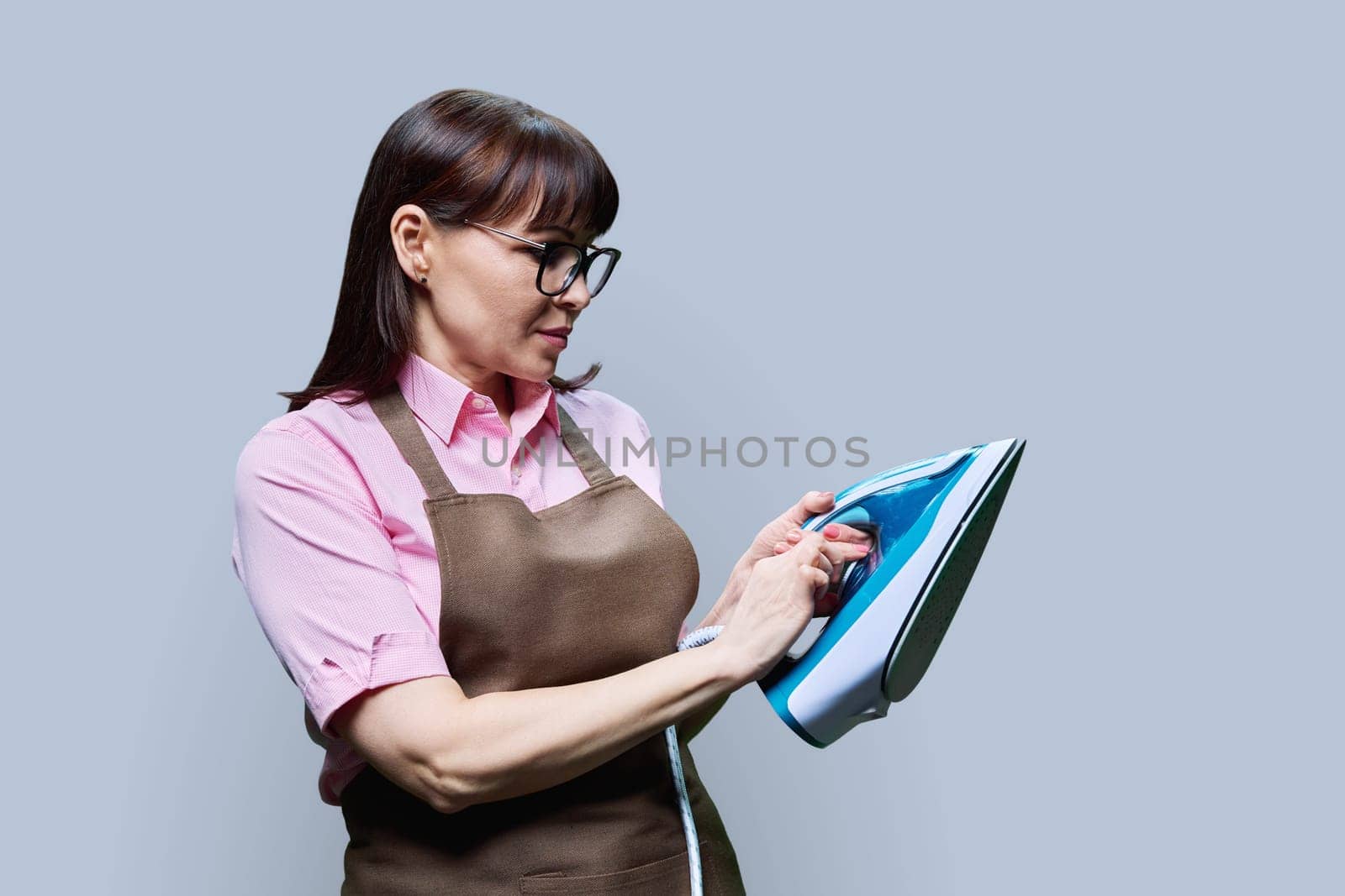 Woman in an apron posing with an iron, on a gray background. Female housewife, dry cleaning laundry worker, lifestyle work staff service concept