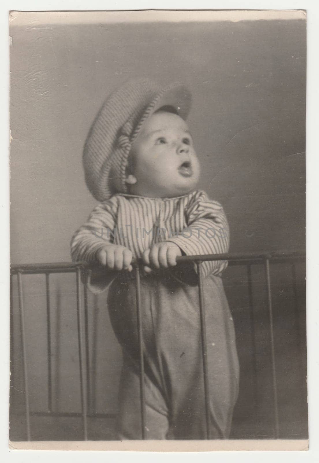 THE CZECHOSLOVAK SOCIALIST REPUBLIC - CIRCA 1960s: Vintage photo shows a small cute boy stands in the cot. He wears a flat cap. Retro black and white photography. Circa 1970s.