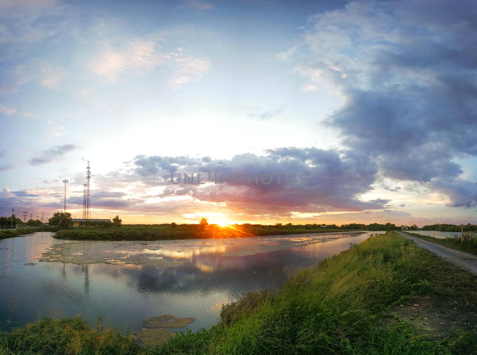 The pond reflects the light from the orange sun. The pool edge is green and yellow grass. The evening sky before the sunset is colorful.