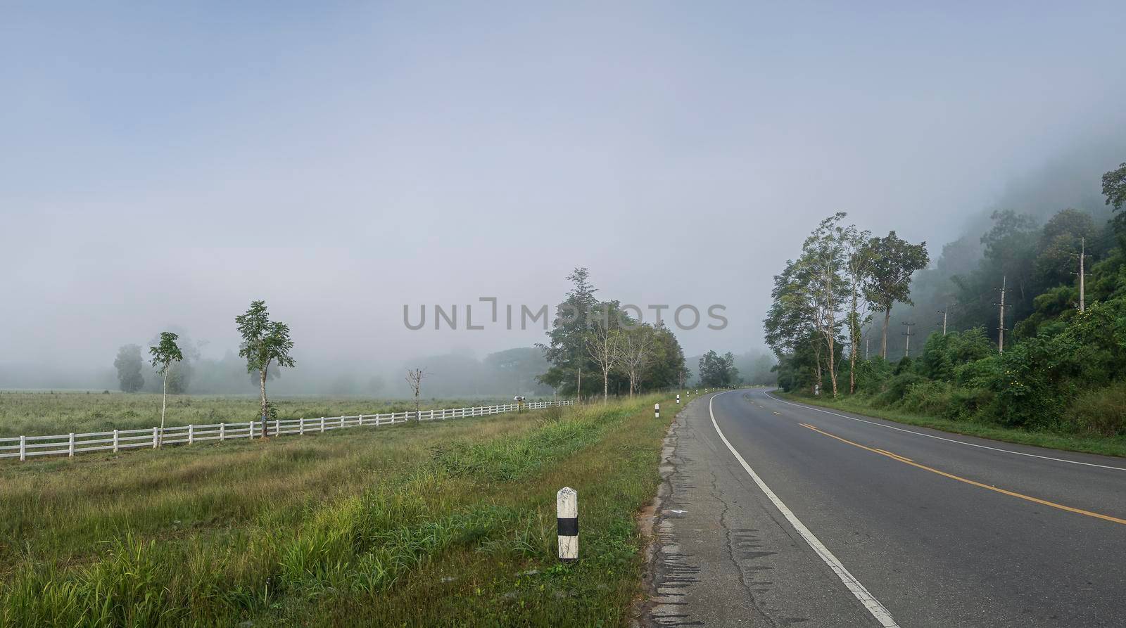 The road between the mountains and the rice fields covered with fog. The road to Pilok Village, Kanchanaburi Province, Thailand.