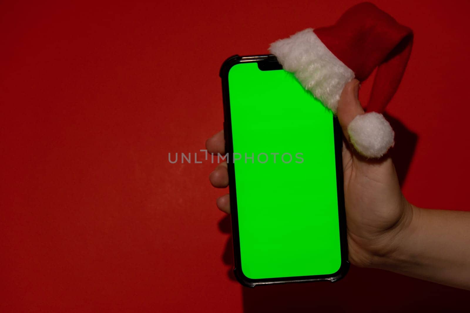 Mobile phone dressed in Santa-Claus red-white hat with chroma key screen against red background. Concept for Christmas or New Years holidays. Female Hand holding Blank cell phone. Digital gadget, technology copyspace wireless wishlist concept. Social media advertisement