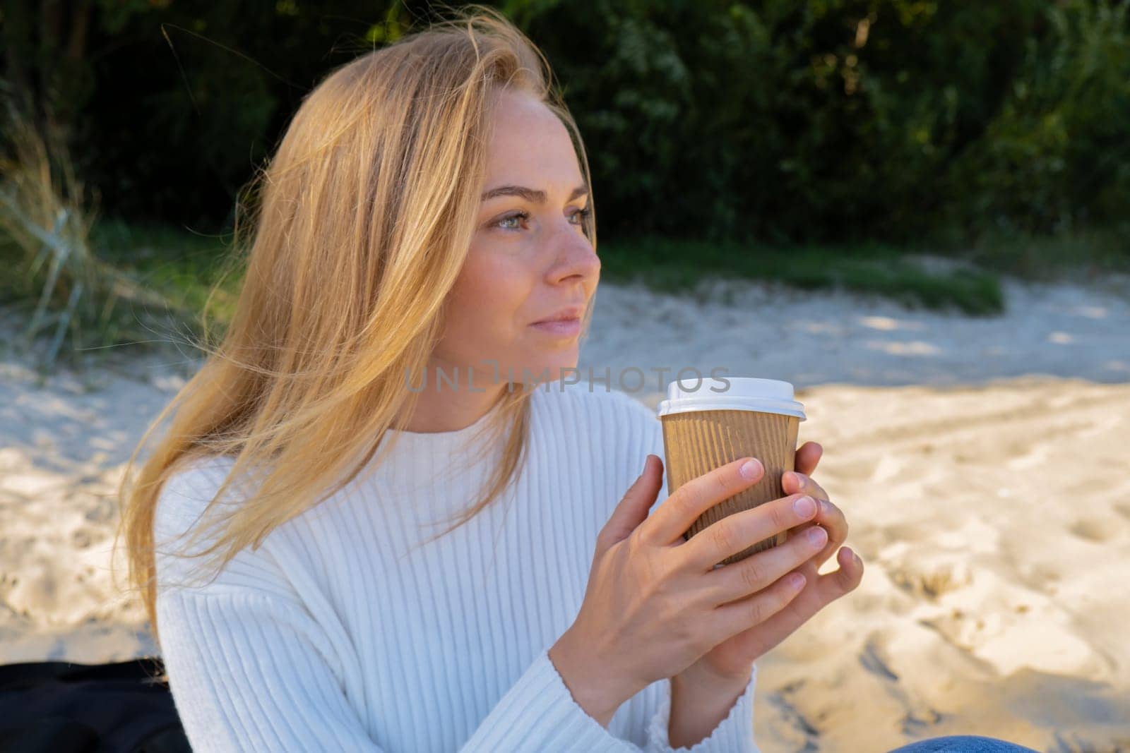 Happy young woman drinking coffee from paper cup takeaway on the beach sea ocean. Wearing wireless headphones doing audio healing sound therapy being mindful Leisure in nature. Wellbeing unity with nature health mindfulness. Enjoy outdoor lifestyle relaxation