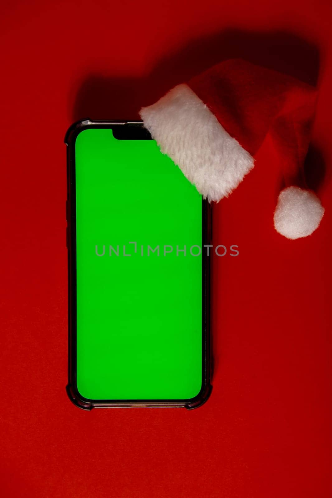 Mobile phone dressed in Santa-Claus red-white hat with chroma key screen against red background. Concept for Christmas or New Years holidays. Blank cell phone. Digital gadget, technology copy space wireless wishlist concept. Social media advertisement