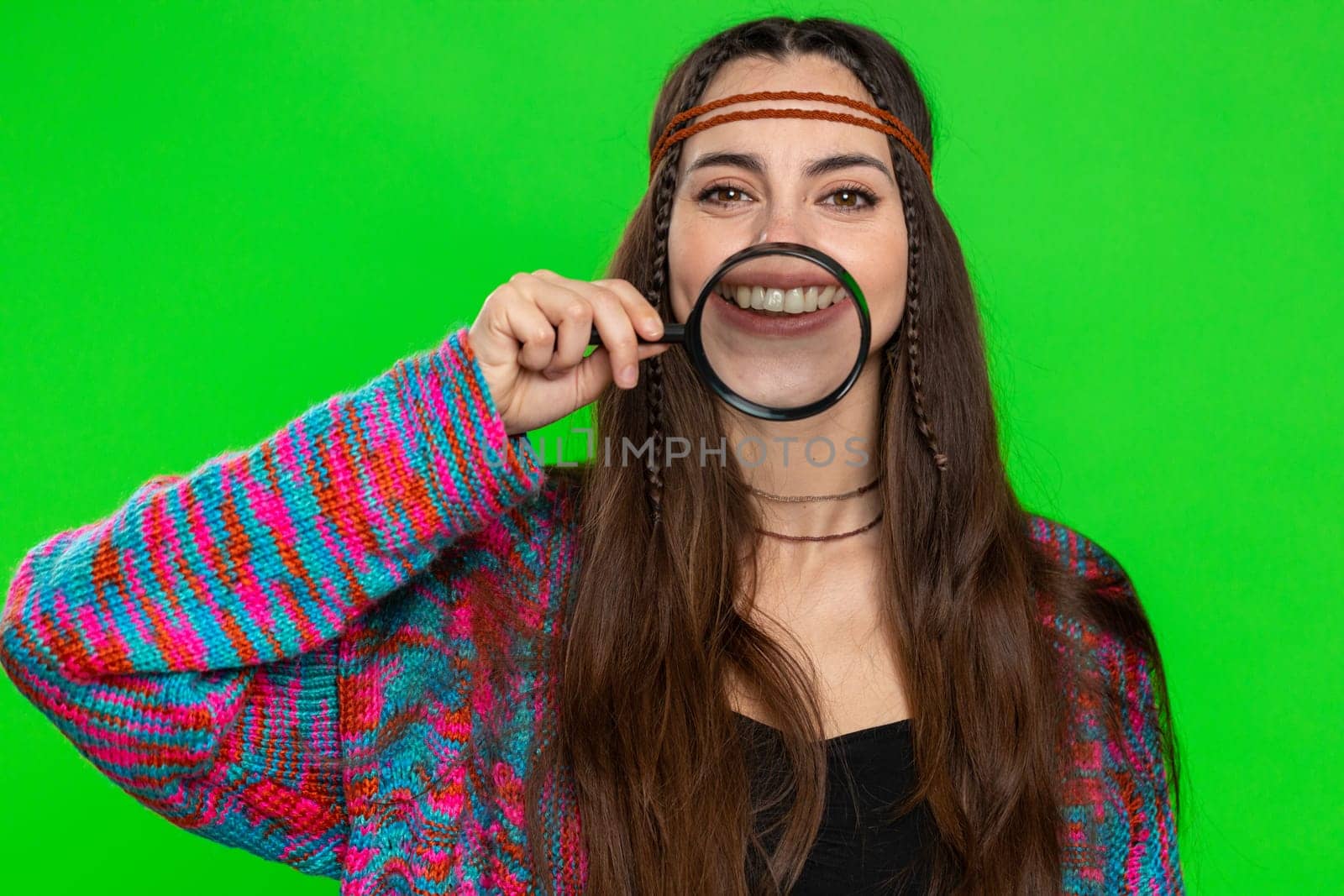 Young woman holding magnifier glass loupe on healthy white teeth, looking at camera with happy expression, showing funny silly face smiling mouth. Pretty hippie girl isolated on chroma key background