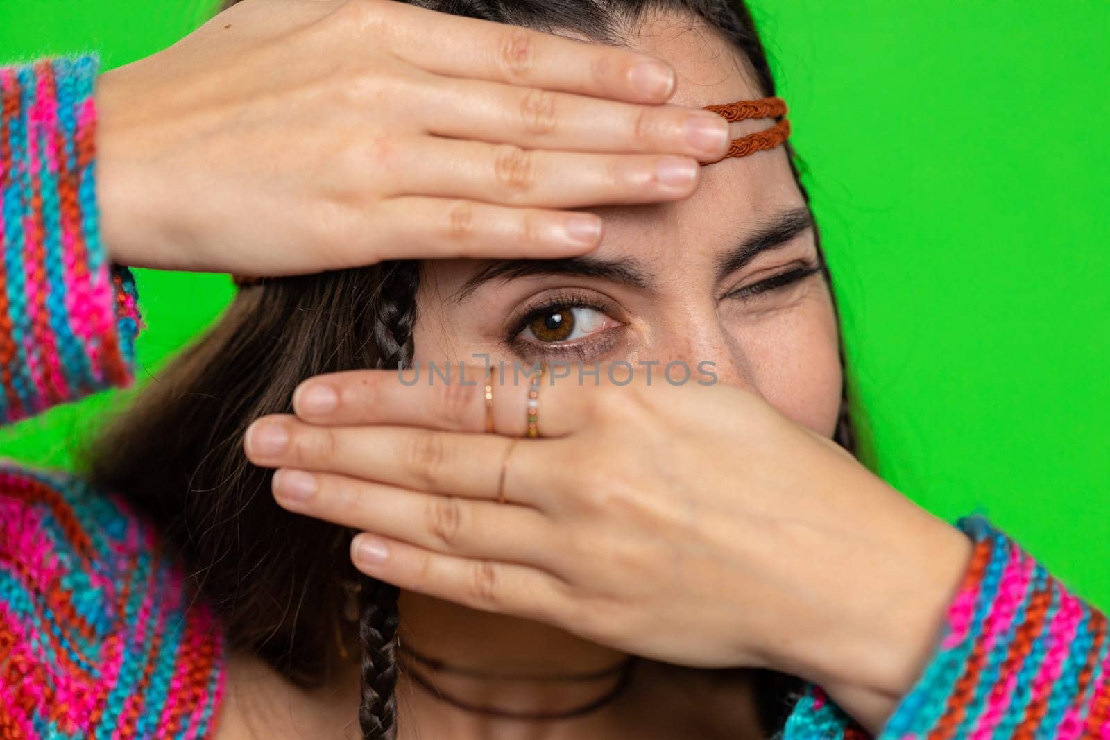 Nosy curious young woman closing eyes with hand and spying through fingers, hiding behind hand fingers, peeping gesture exploring way seeking something in distance. Girl on green chroma key background