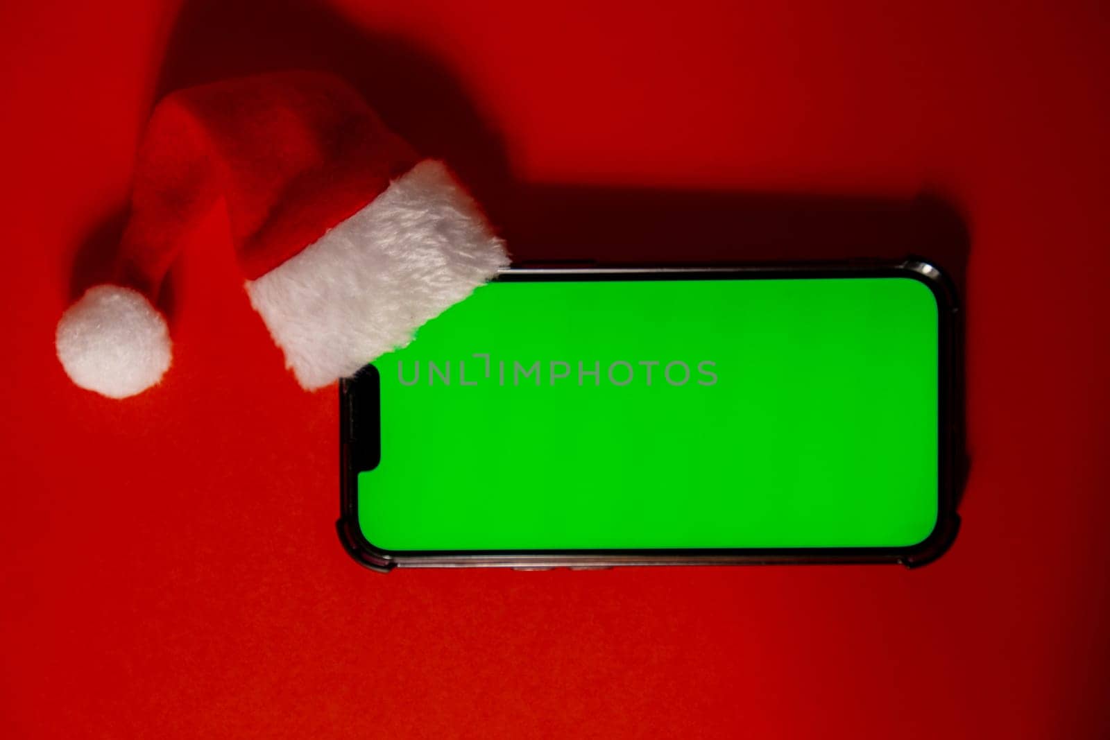 Mobile phone dressed in Santa-Claus red-white hat with chroma key screen against red background. Concept for Christmas or New Years holidays. Blank cell phone. Digital gadget, technology copy space wireless wishlist concept. Social media advertisement