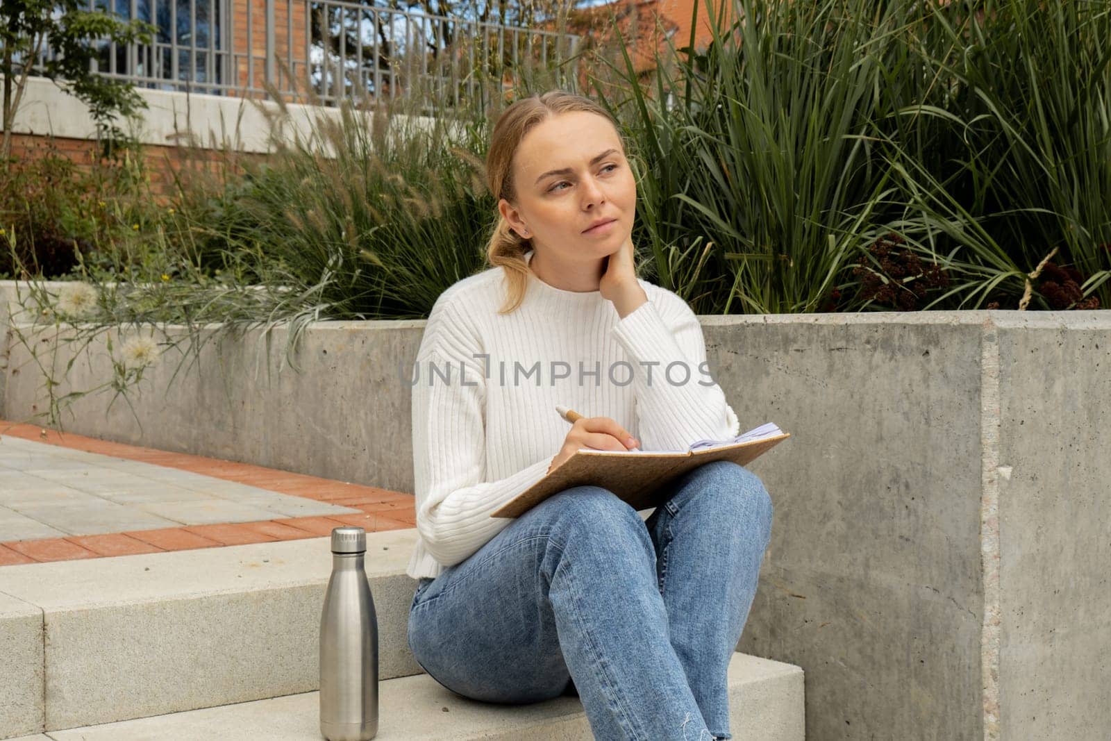Young student study with notebook in park. Drinking water, hot tea or coffee from reusable metal bottle. Writing gratitude journal self reflection self discovery by anna_stasiia