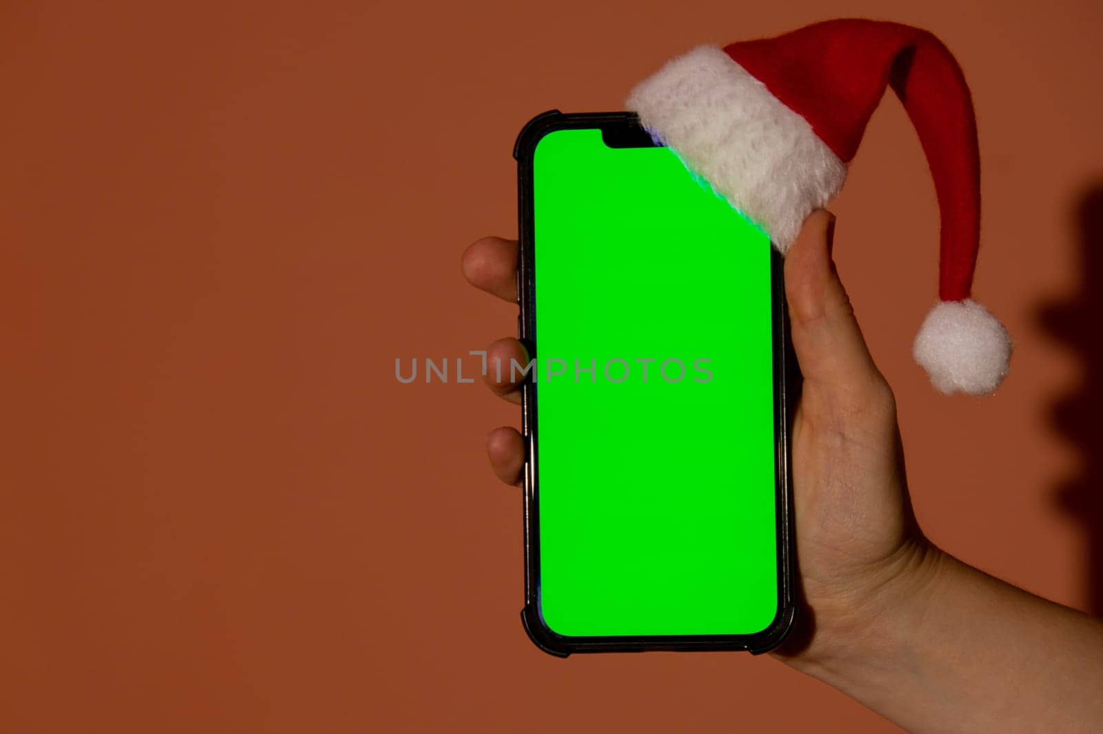 Mobile phone dressed in Santa-Claus red-white hat with chroma key screen against red background. Concept for Christmas or New Years holidays. Female Hand holding Blank cell phone. Digital gadget, technology copyspace wireless wishlist concept. Social media advertisement