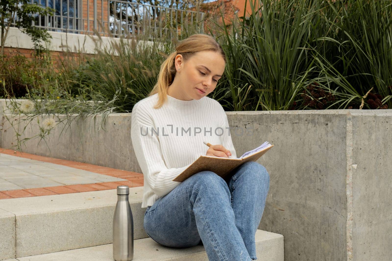 Young student study with notebook in park. Drinking water, hot tea or coffee from reusable metal bottle. Writing gratitude journal self reflection self discovery by anna_stasiia