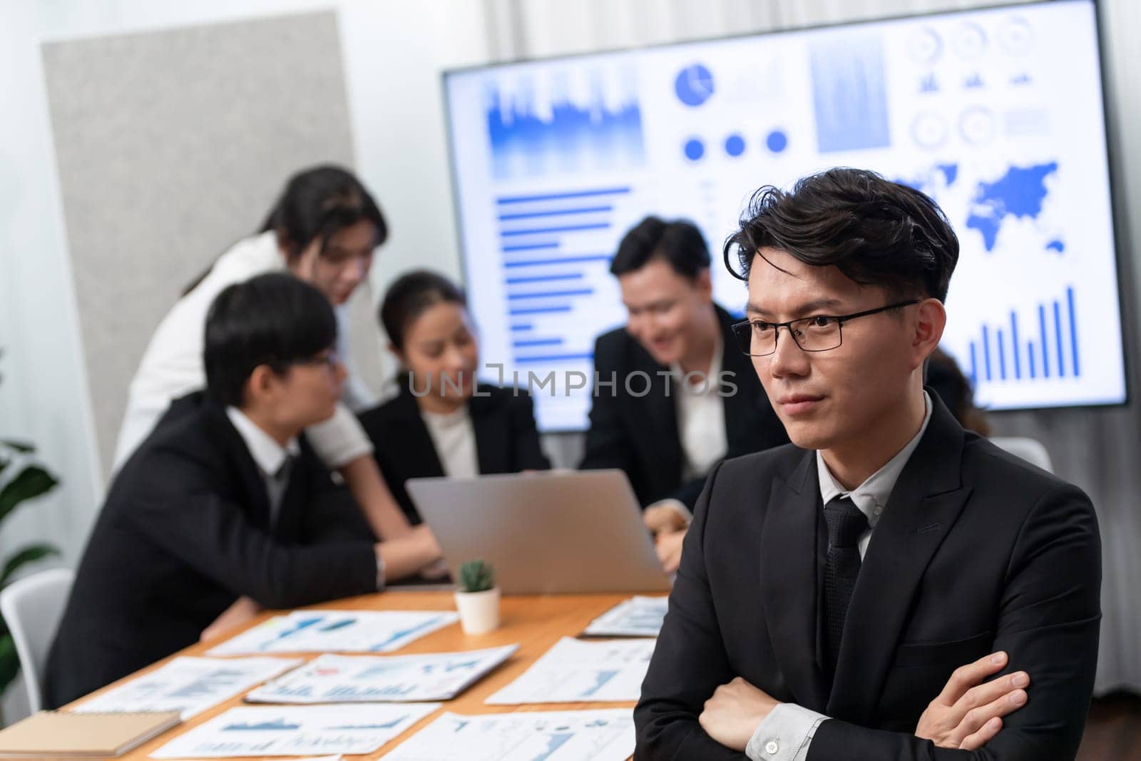 Focus portrait of successful confident male manager or executive in business wear with blurred background of businesspeople, colleagues working with financial report papers in office of harmony.