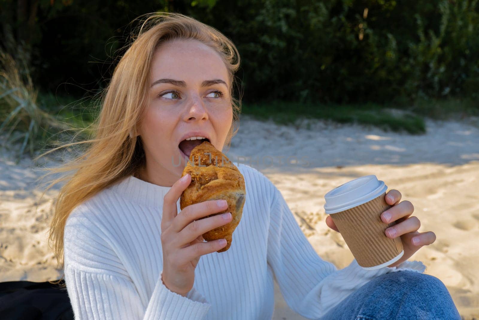 Happy young woman drinking coffee from paper cup takeaway food eating croissant on the beach sea ocean. Enjoy autumn cloudy day. Leisure in nature. Calm and relaxation concept. Traveling concept Wellbeing unity with nature health mindfulness. Enjoy outdoor lifestyle relaxation