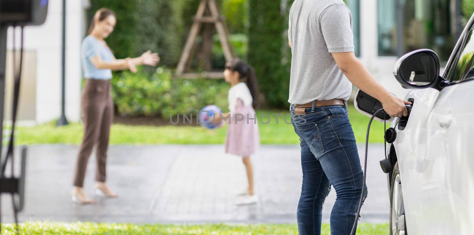 Focus image of progressive man charging electric car from home charging station with blur mother and daughter playing together in the background.