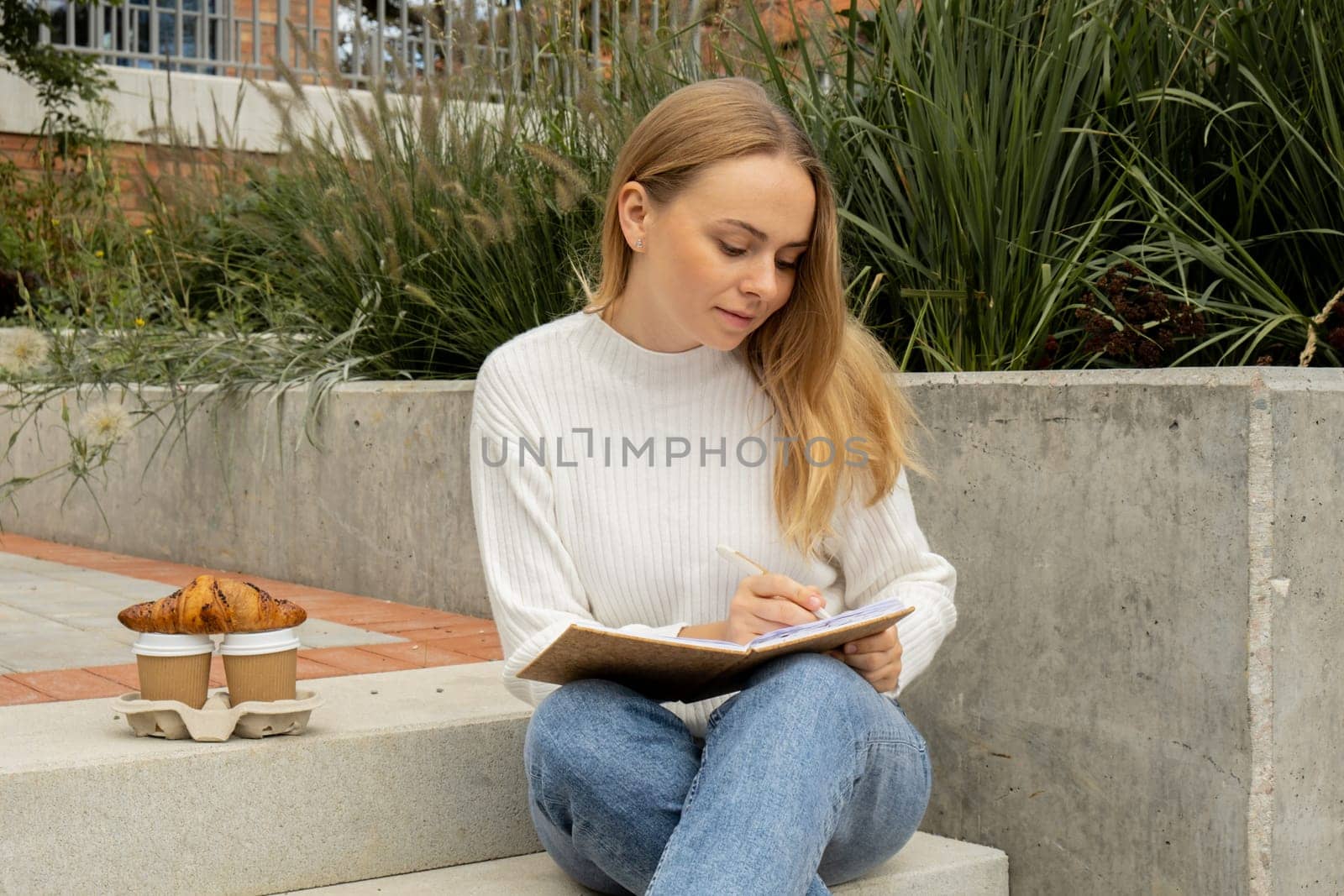 Young student study with notebook in park. Drinking coffee and eat croissants. Writing gratitude journal self reflection self discovery Outdoors warm autumn seashore.