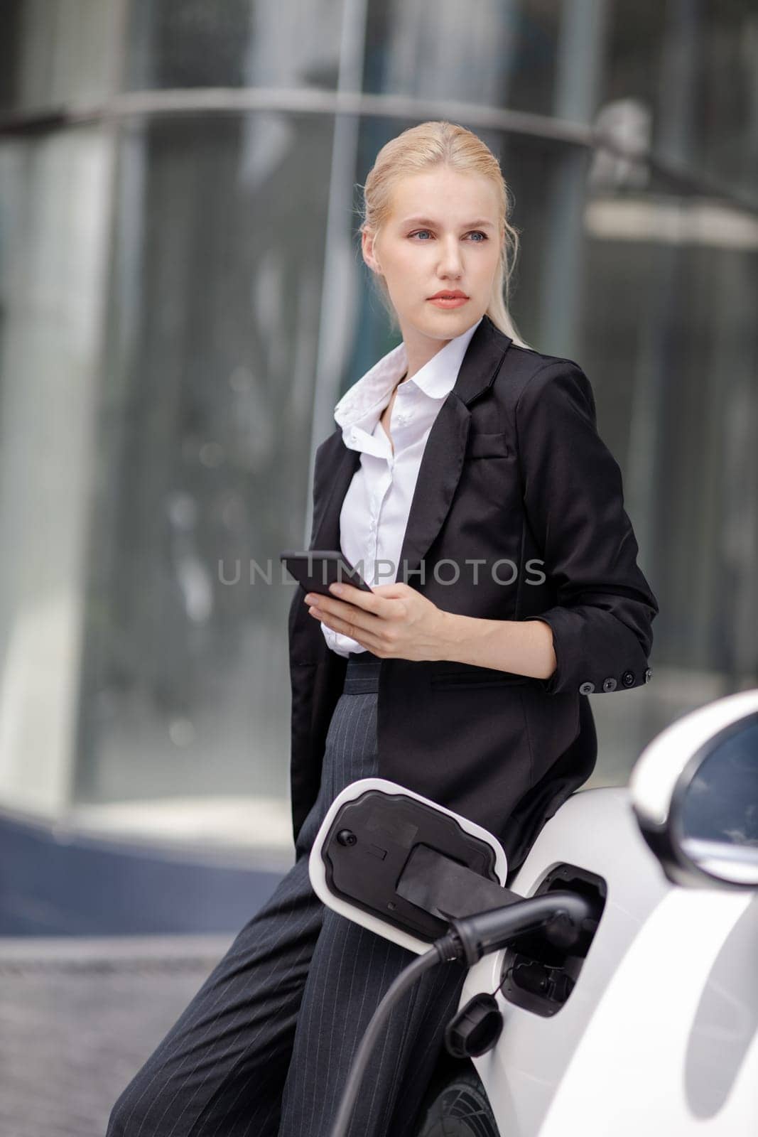 Progressive businesswoman with mockup smartphone with EV at charging station. by biancoblue