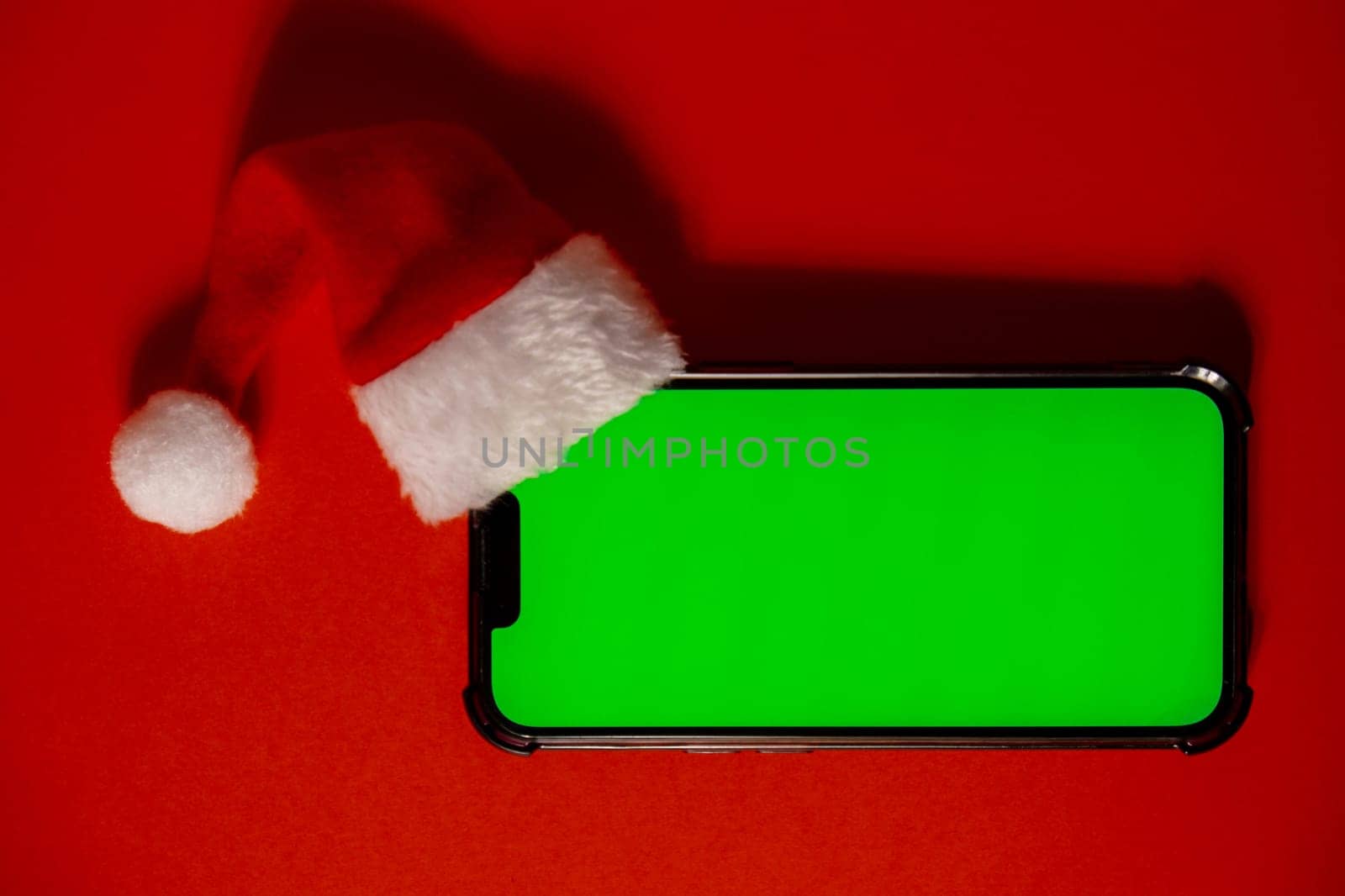 Mobile phone dressed in Santa-Claus red-white hat with chroma key screen against red background. Concept for Christmas or New Years holidays. Blank cell phone. Digital gadget, technology copy space wireless wishlist concept. Social media advertisement