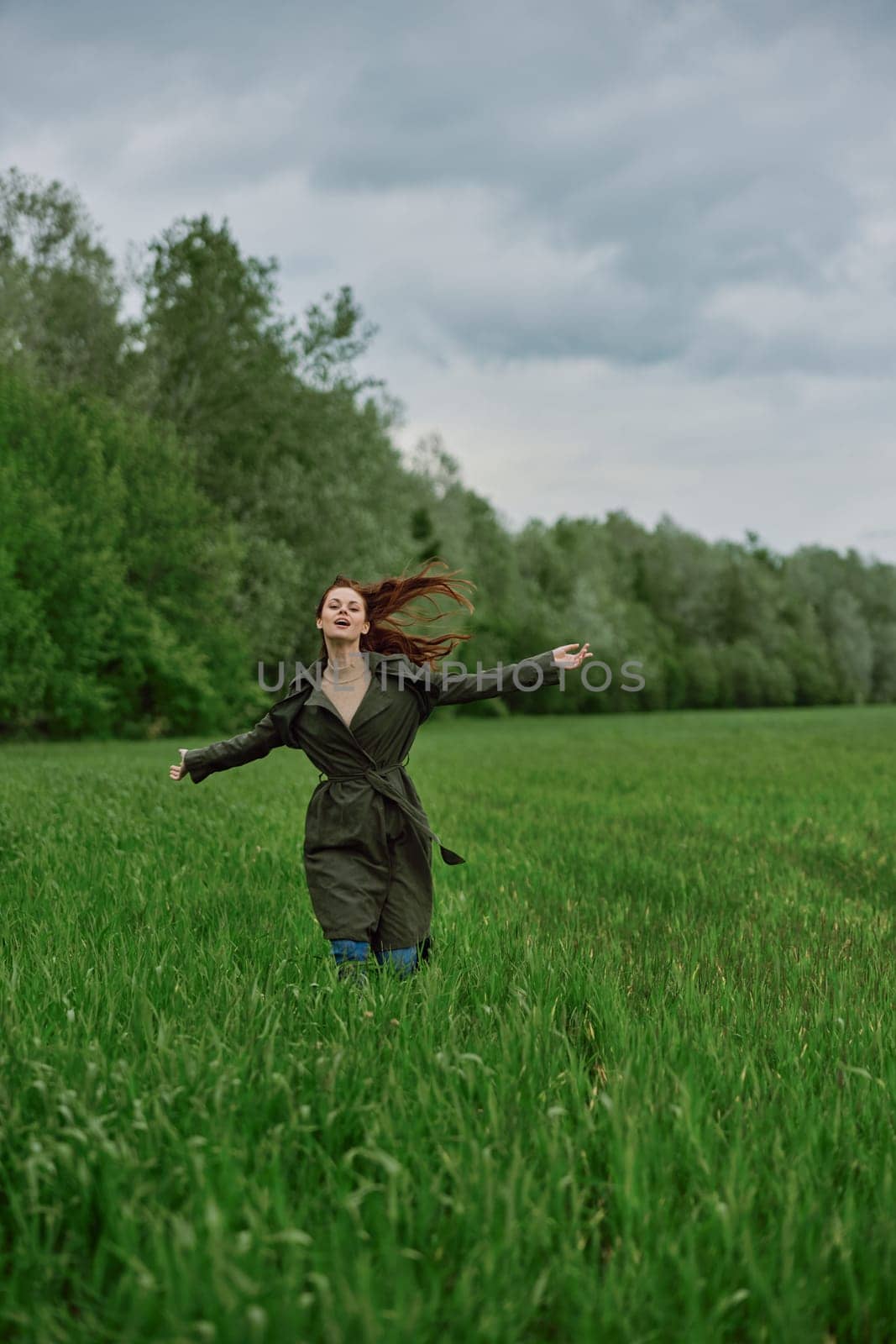 a beautiful woman in a long raincoat runs across a field in high grass in spring in cloudy weather. High quality photo