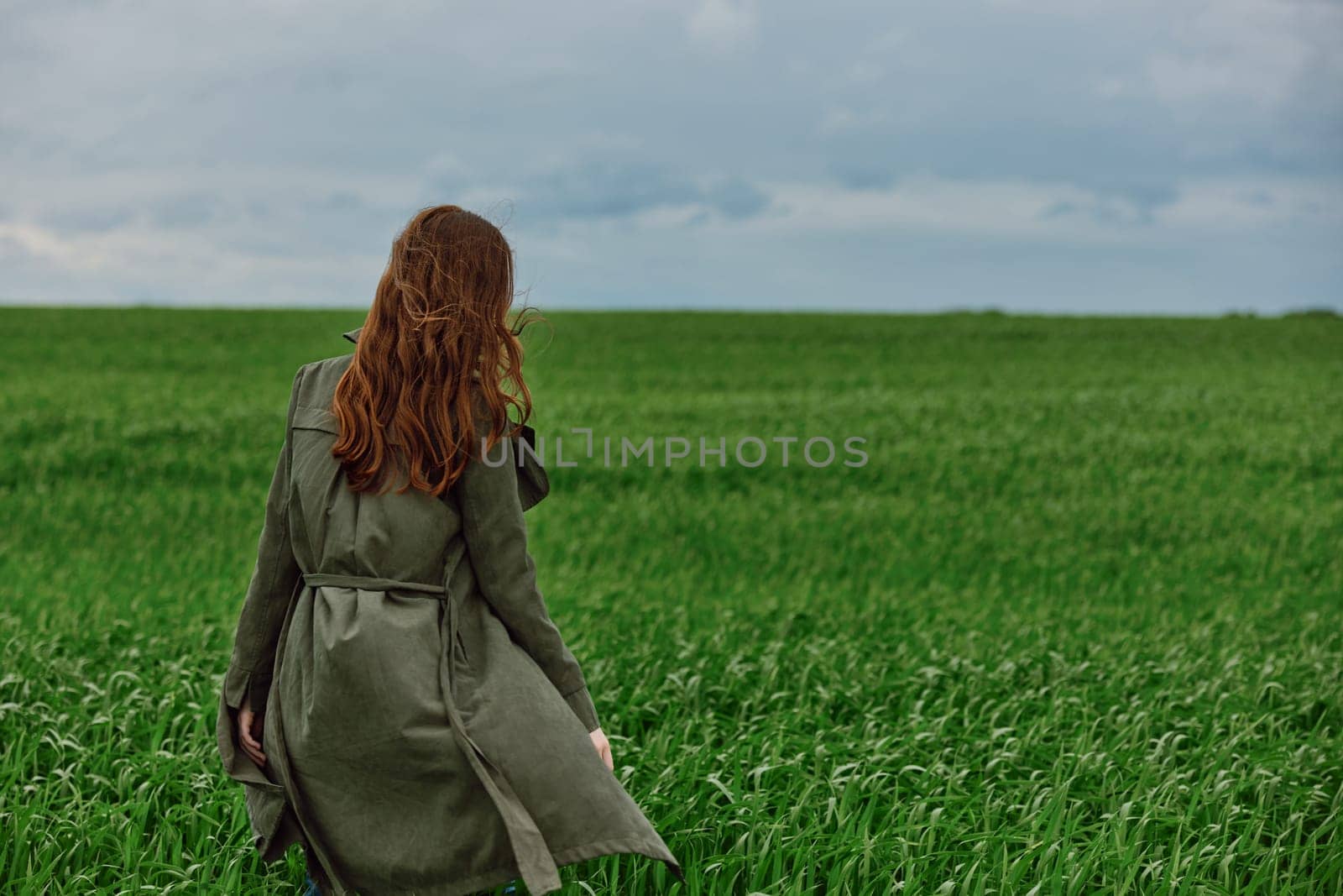 a red-haired woman in a long raincoat stands in a green field in cloudy weather in a strong wind by Vichizh