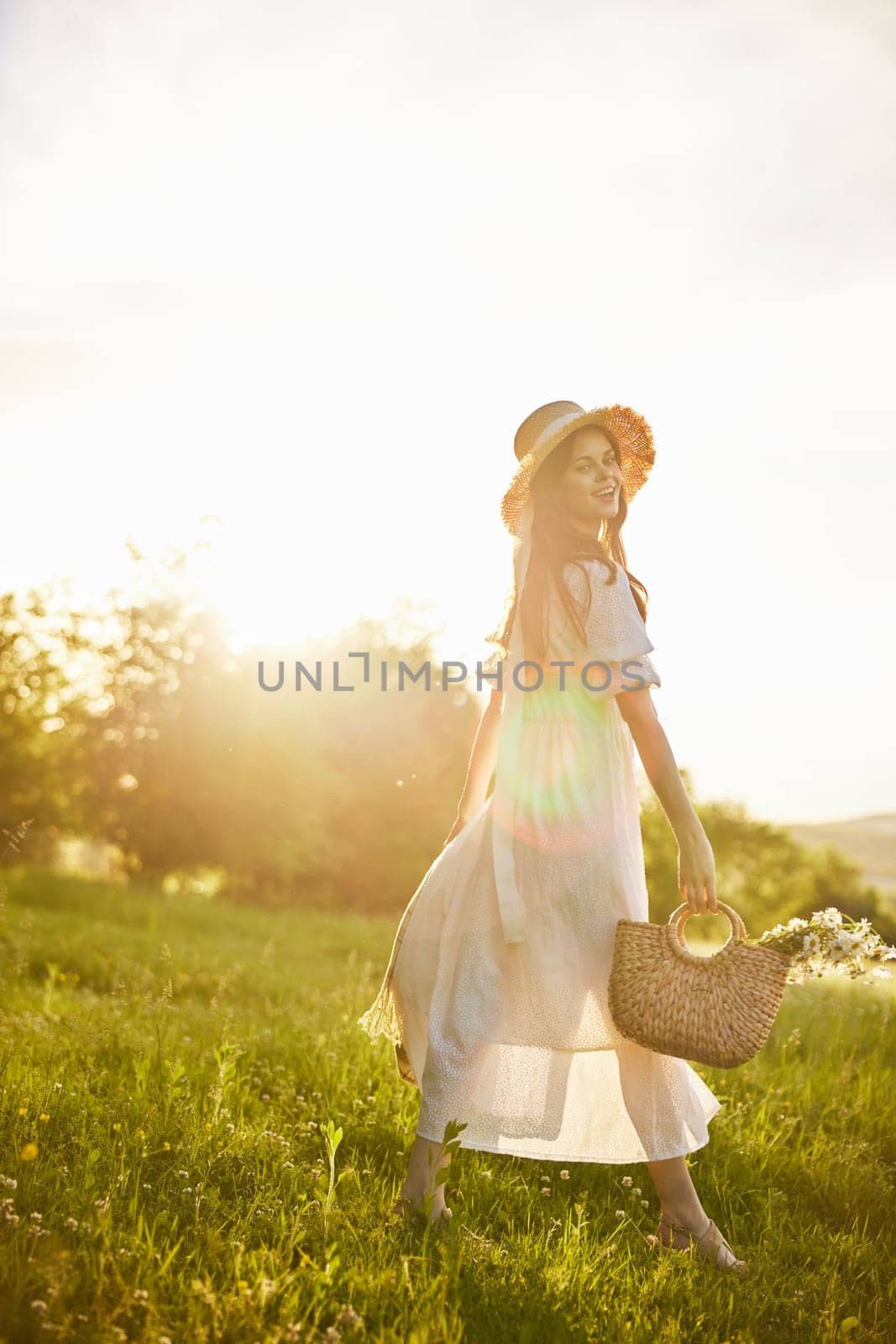 a woman in a long light dress walks through the countryside in a hat and with a basket in her hands in the rays of the setting sun enjoying nature by Vichizh