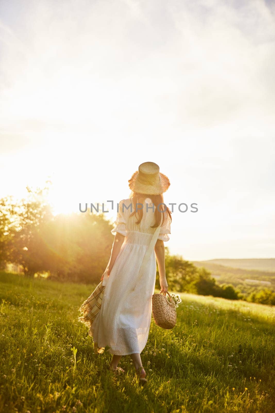 a woman in a long light dress walks through the countryside in a hat and with a basket in her hands in the rays of the setting sun enjoying nature. Photo from the back. High quality photo