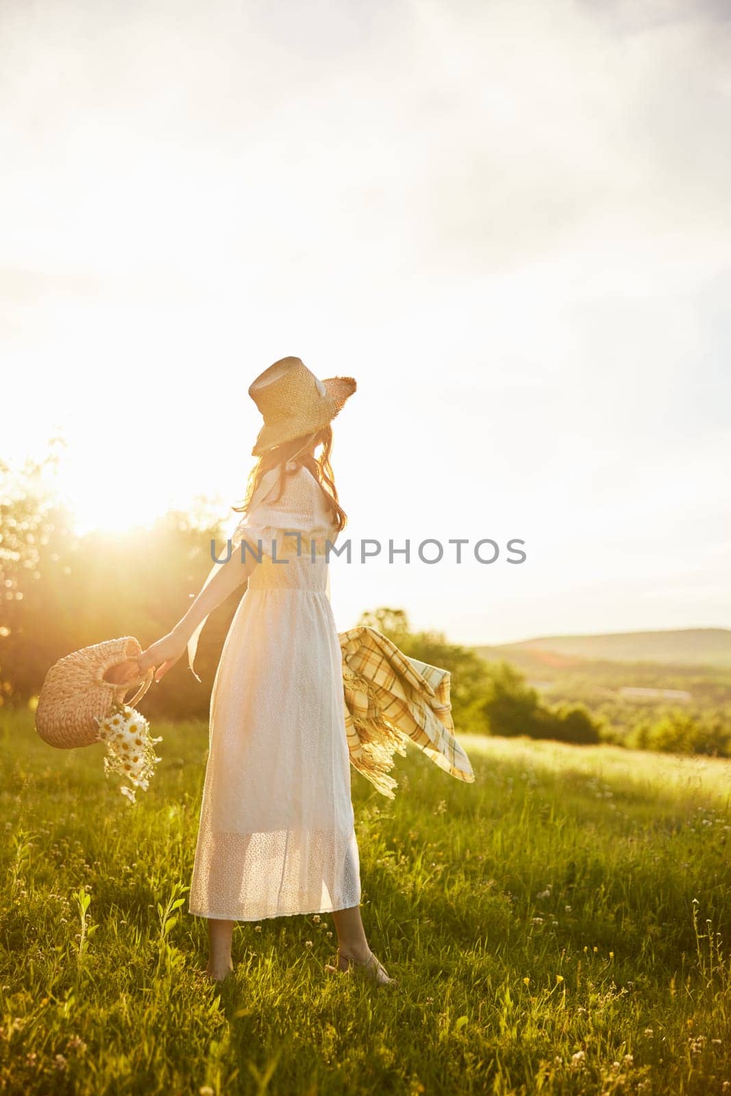 a woman in a long light dress walks through the countryside in a hat and with a basket in her hands in the rays of the setting sun enjoying nature by Vichizh