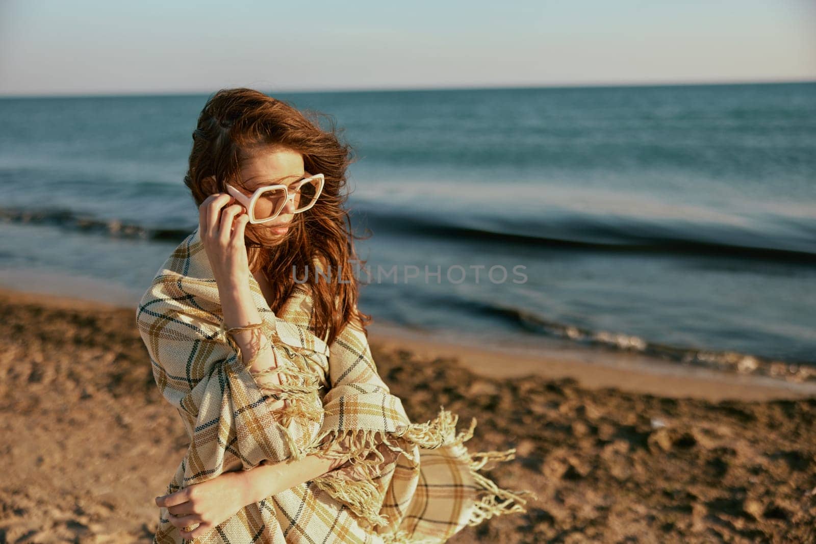 a woman wrapped in a plaid walking along the sea coast straightens her sunglasses. High quality photo