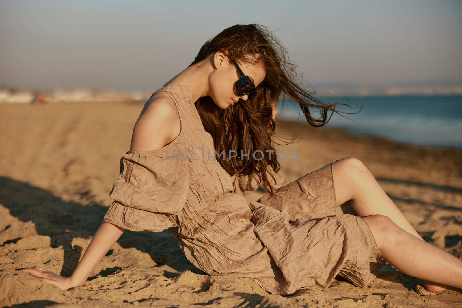 red-haired woman in dark sunglasses and sand-colored dress on the beach enjoying the sunset by Vichizh