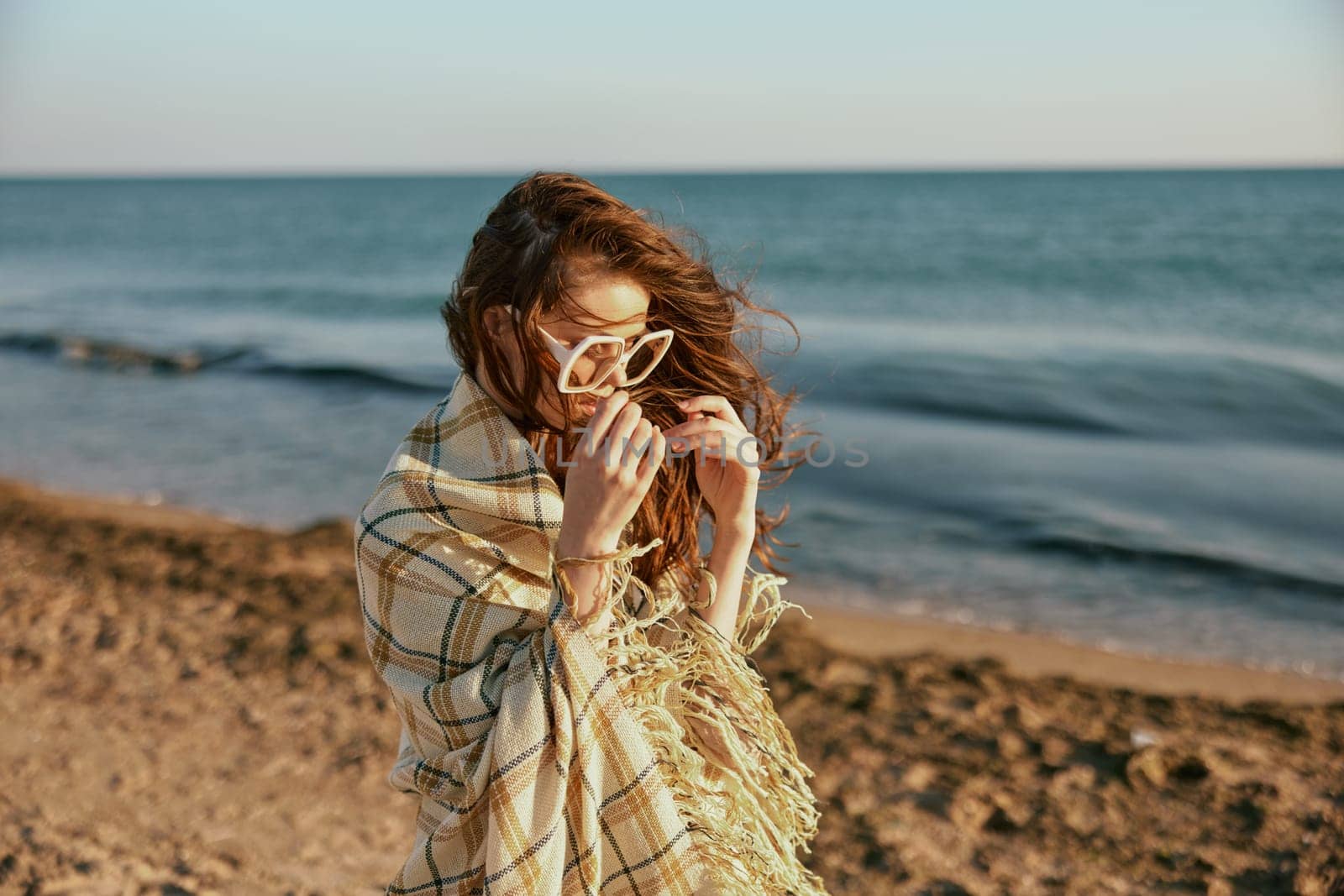 a woman wrapped in a plaid walking along the sea coast straightens her sunglasses by Vichizh