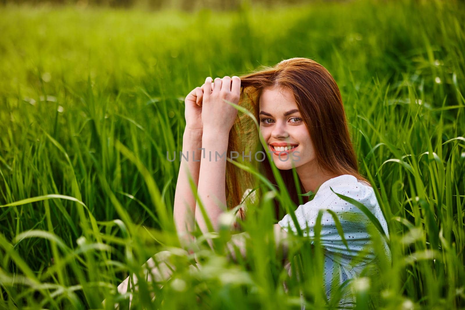 portrait of a beautiful, happy woman sitting in the grass in a light dress and smiling at the camera by Vichizh