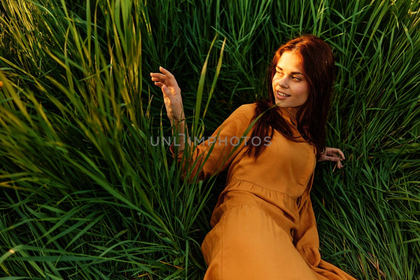 a happy woman is lying in the tall green grass in a long orange dress and smiling pleasantly looks away holding the leaves with her hand, enjoying nature. High quality photo