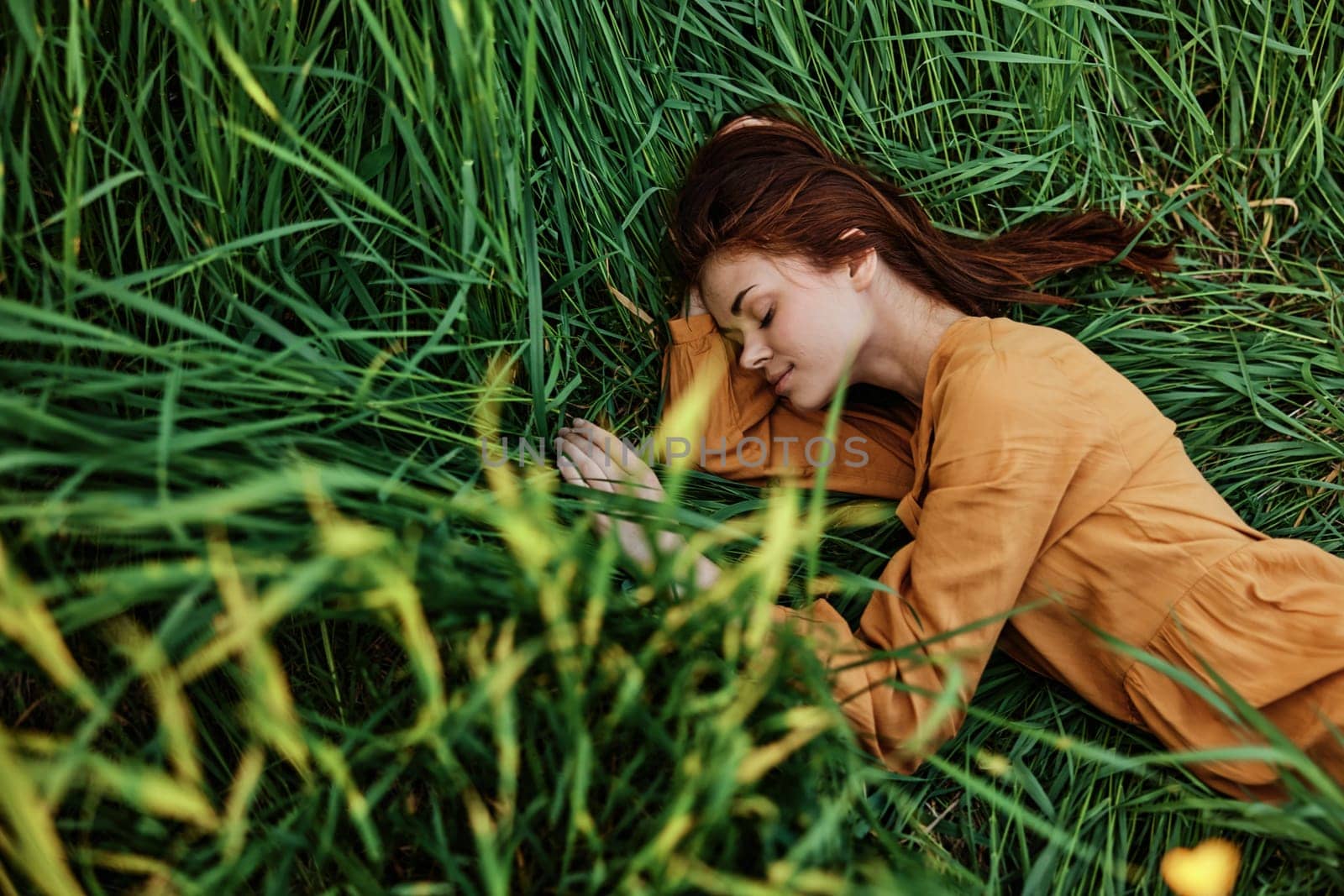 a close horizontal photo of a pleasant woman in a long orange dress resting lying in the tall grass with her eyes closed in sunny weather at sunset with her arms outstretched. Street photography, the theme of privacy with nature. High quality photo