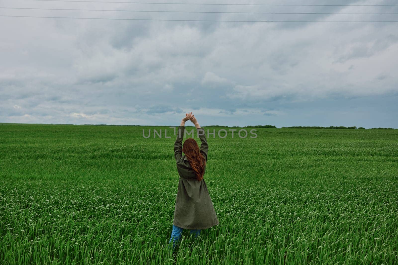a woman in a long coat stands in a green field on a cloudy day with her hands up to the sky by Vichizh