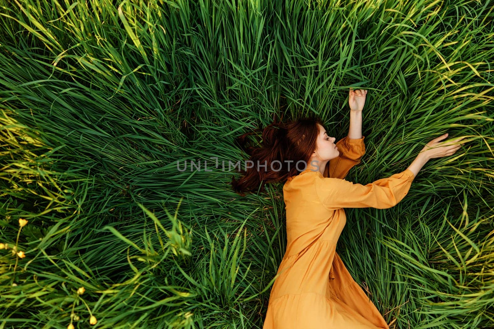 an elegant woman lies in the tall green grass in a long summer orange dress and relaxed posing with her hands enjoying nature and sunny weather with her eyes closed. High quality photo