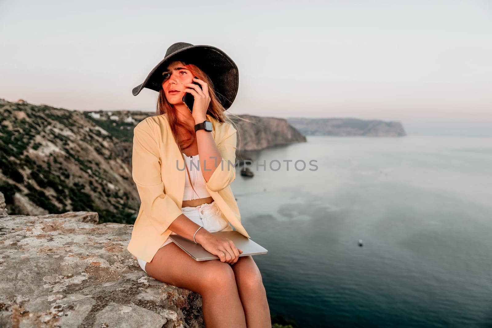 Successful business woman in yellow hat working on laptop by the sea. Pretty lady typing on computer at summer day outdoors. Freelance, travel and holidays concept.