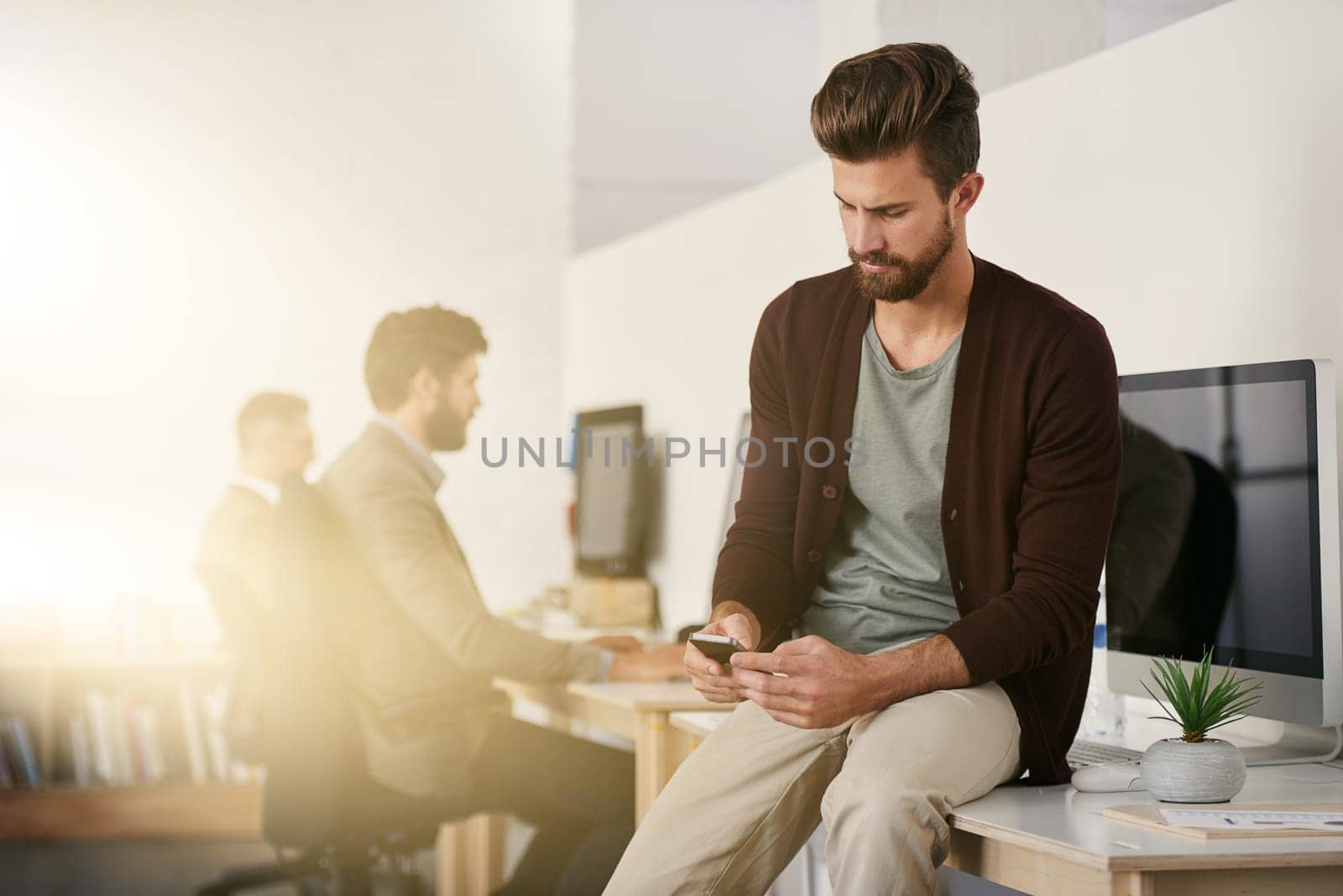 Confident and creative. a young designer using his cellphone with his colleagues blurred in the background. by YuriArcurs