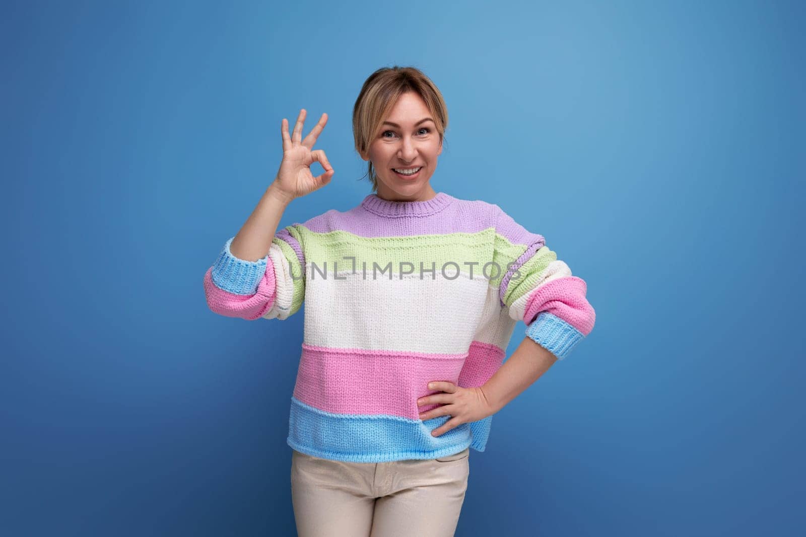 attractive blond young woman in pastel colored sweater smiling showing class on blue background.