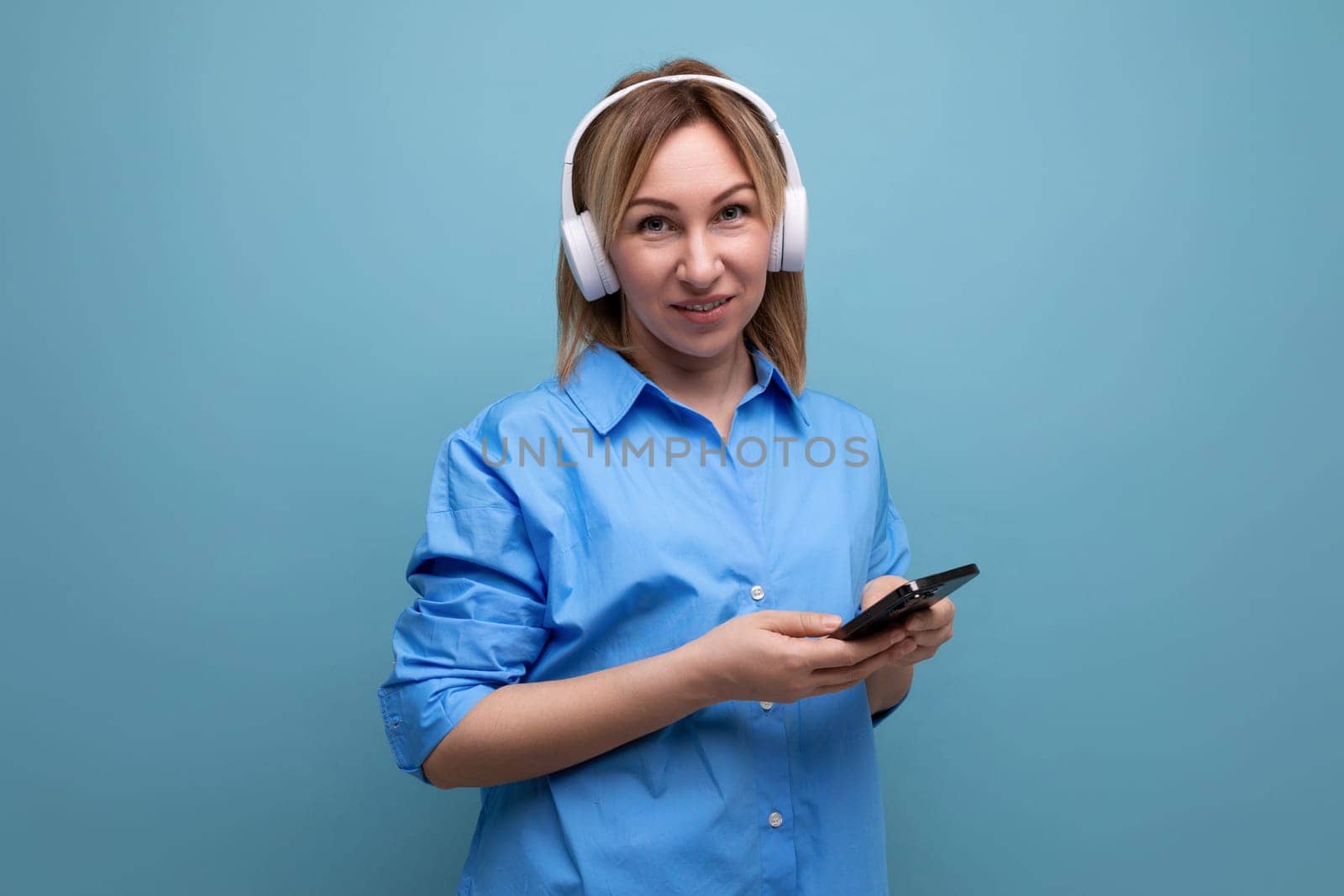 widescreen photo of a pensive european girl in a casual shirt listening to music in big white headphones on a blue isolated background by TRMK
