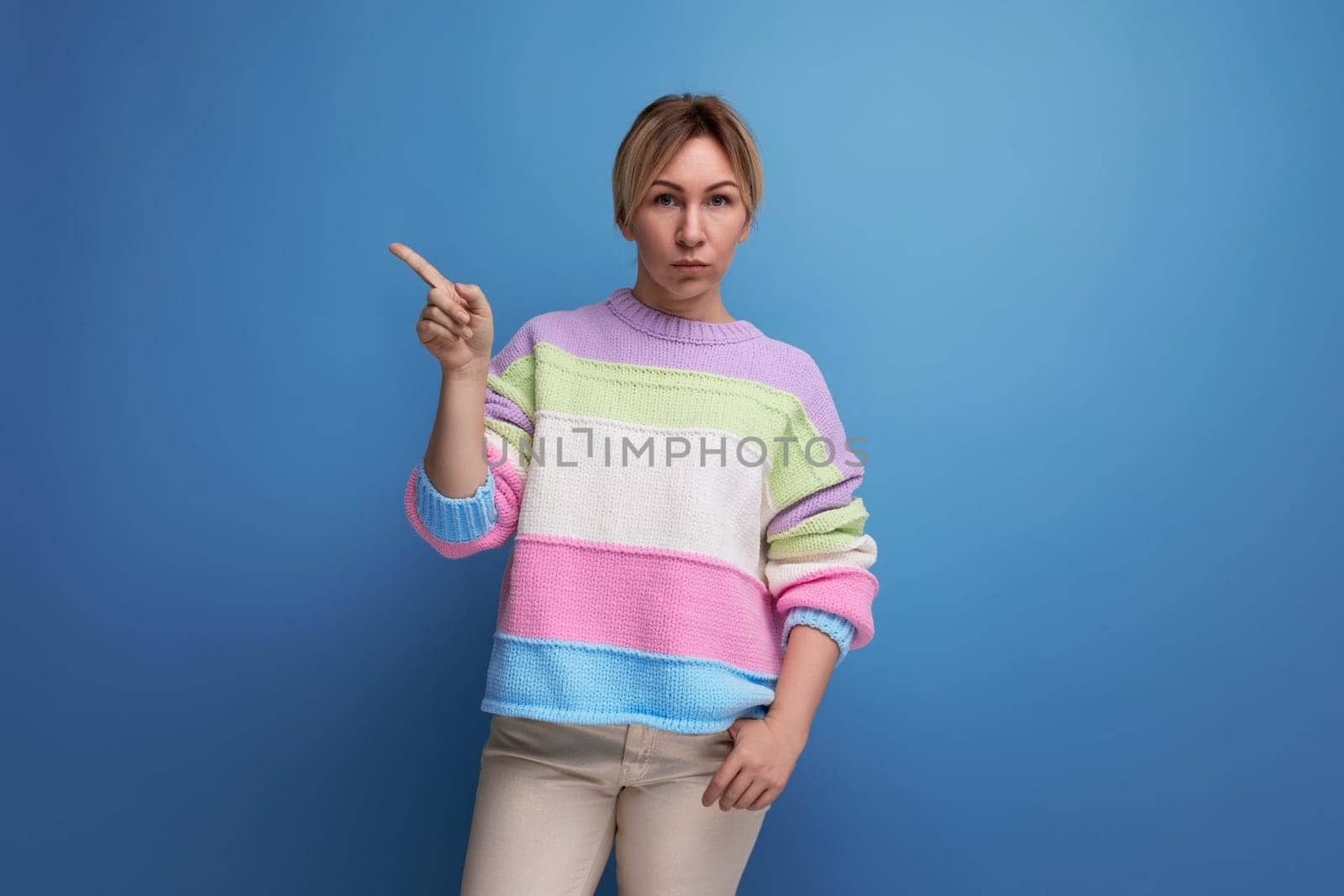 serious blond young woman in a striped sweater shows her hand in on a blue background by TRMK