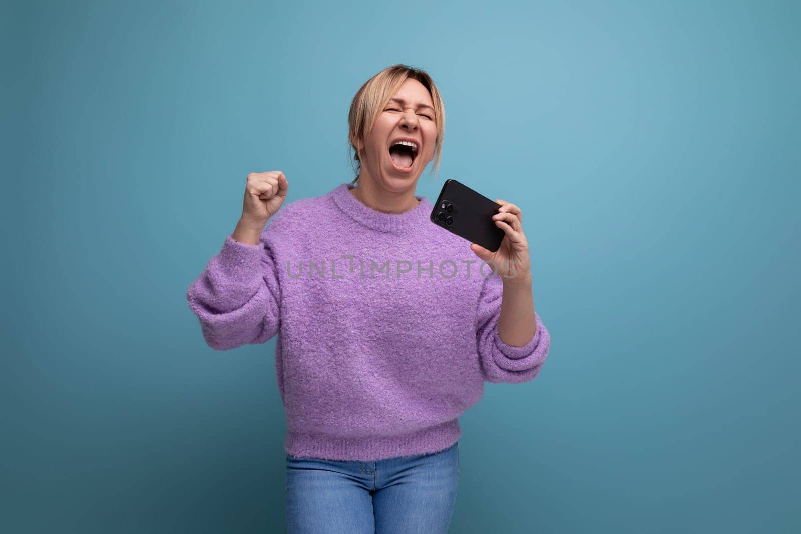 energetic blond young woman consultant in purple hoodie holding smartphone with mockup and singing on blue background with copy space.