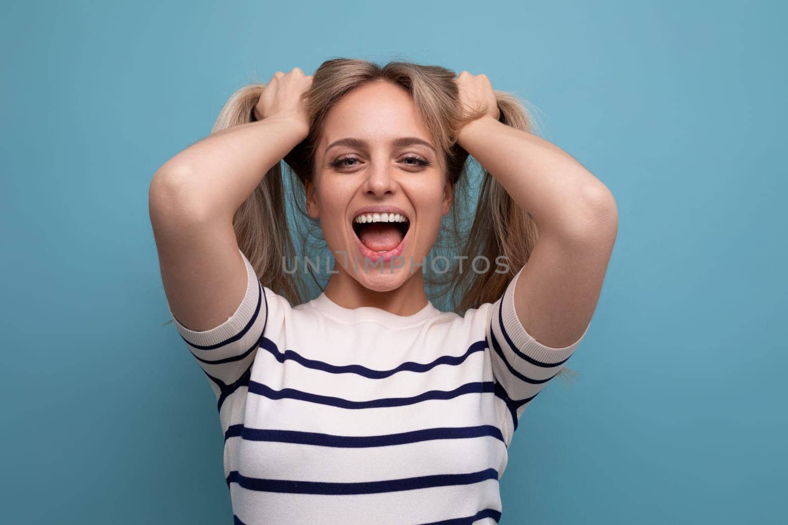 cheerful blond young woman holds her hair in her hands and laughs cutely on a blue background.