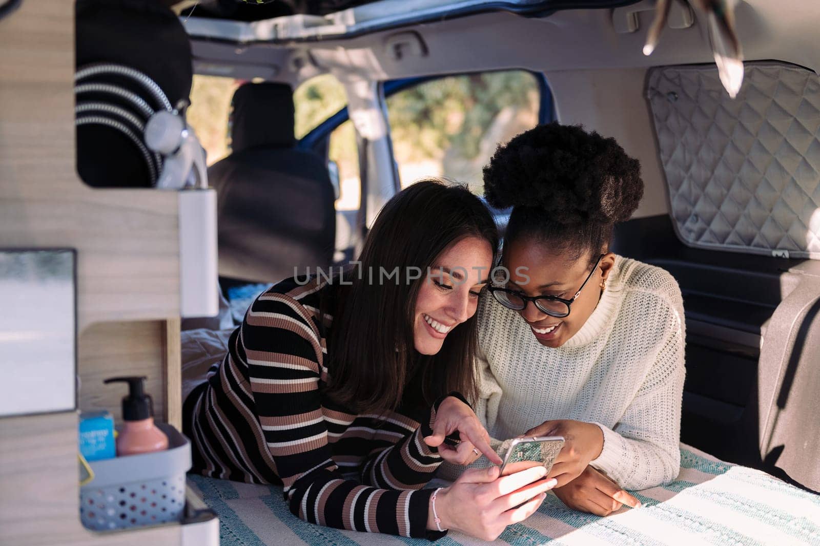 two young women smiling happy and enjoying with mobile phone in camper van, concept of nomadic lifestyle and van life relaxation