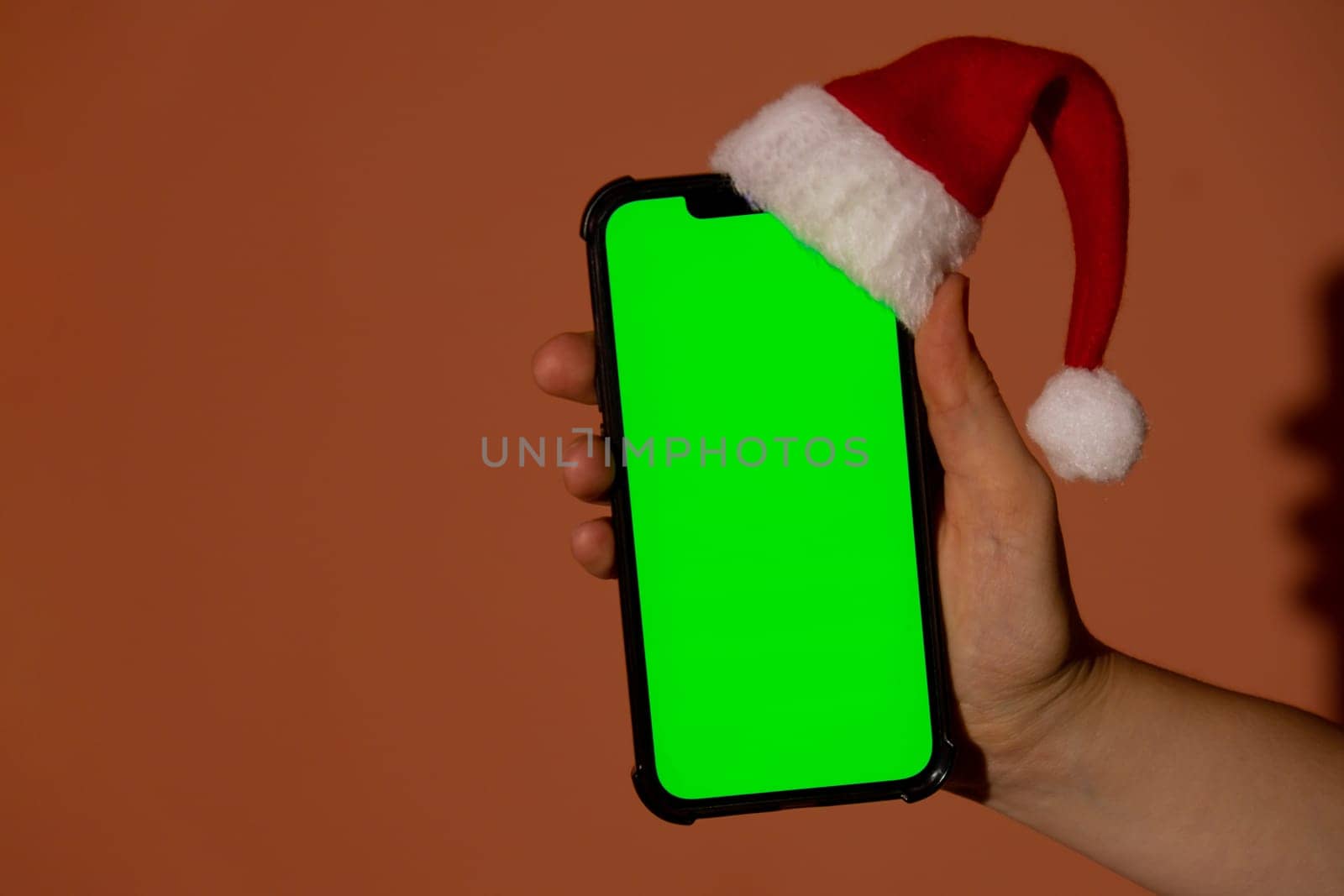 Mobile phone dressed in Santa-Claus red-white hat with chroma key screen against red background. Concept for Christmas or New Years holidays. Female Hand holding Blank cell phone. Digital gadget, technology copyspace wireless wishlist concept. Social media advertisement