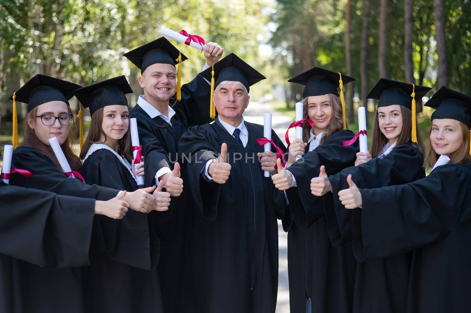 A group of graduates in robes give a thumbs up outdoors. Elderly student