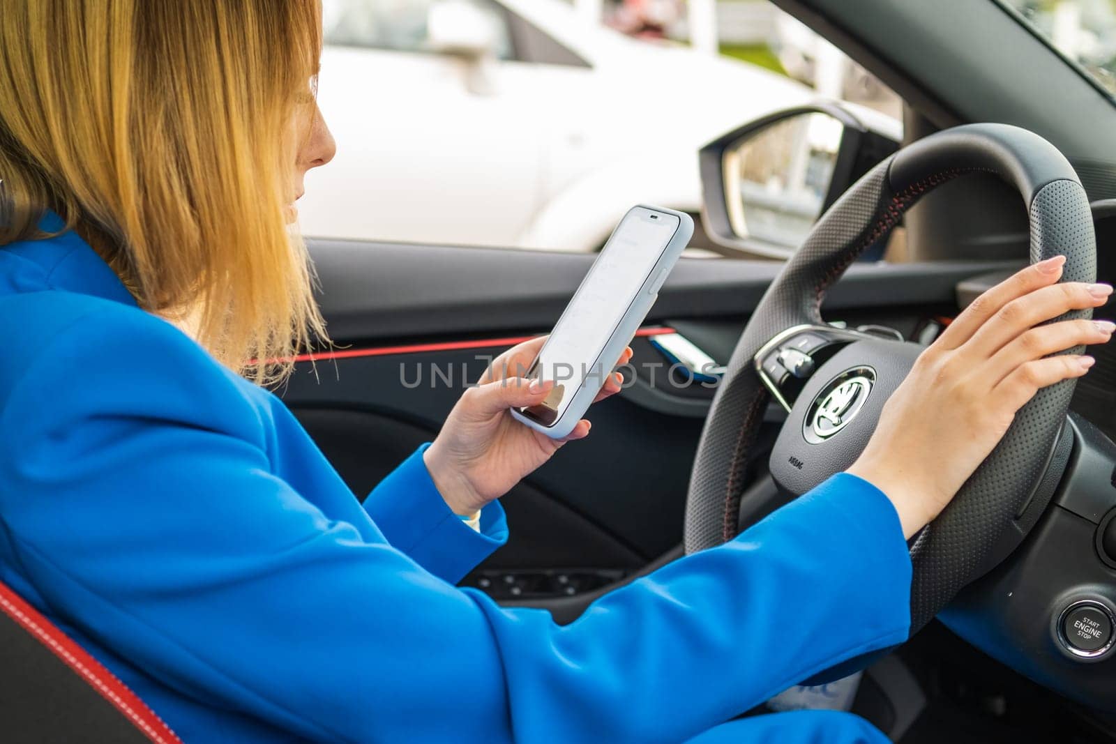 Young smiling woman in the blue suit sitting in the car and texting on her mobile phone